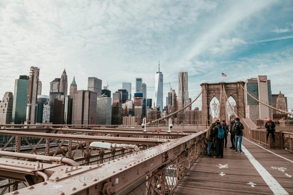 A view from the pedestrian walkway of the Brooklyn Bridge showing tourists strolling along the wooden path, with a stone bridge tower ahead and the skyscrapers of the Lower Manhattan skyline—including One World Trade Center—dominating the background under a cloudy sky.